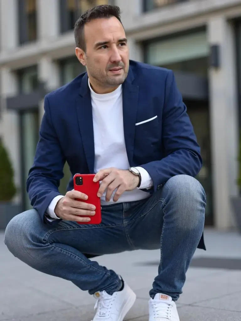 Christian Gottschling hockt mit rotem Smartphone in der Hand vor einer modernen Gebäudefassade in der Stadt, Blick zur Seite