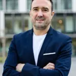 Christian Gottschling mit verschränkten Armen vor der Glasfassade eines modernen Bürogebäudes, blaues Sakko und weißes Shirt