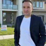 Christian Gottschling in dunklem Sakko und Jeans steht bei Sonnenschein vor einem modernen Wohnhaus mit Balkon und Rasen